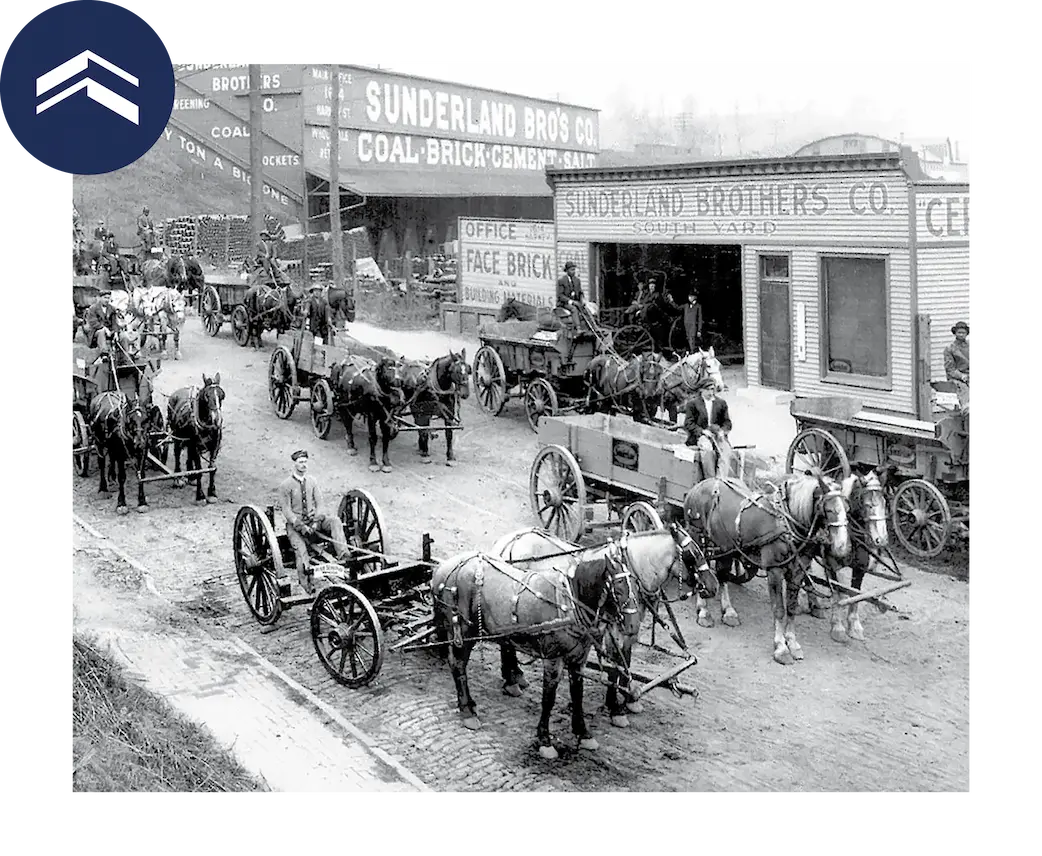 Historic black-and-white photograph of horse-drawn wagons lined up outside the Sunderland Brothers Coal, Brick, and Cement yard, with workers standing beside carts on a busy industrial street.