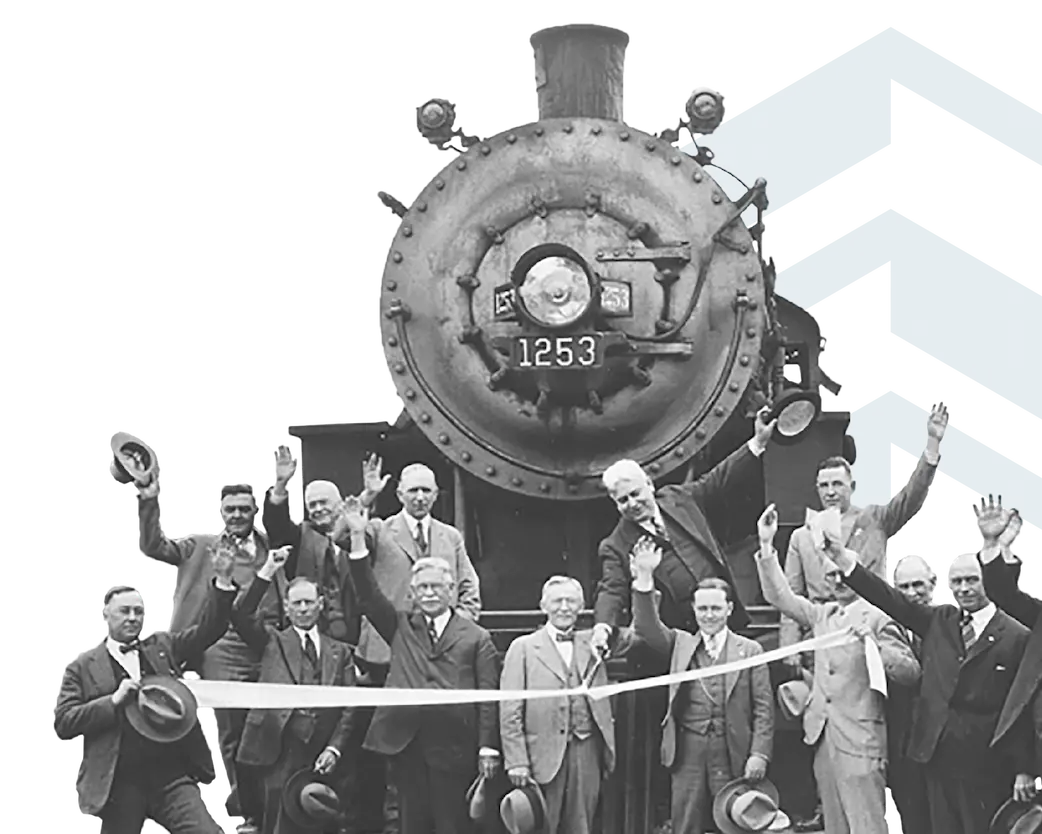 Historic black-and-white photograph of men in suits waving in front of a steam locomotive numbered 1253, with a ribbon stretched across the foreground, marking a ceremonial event.