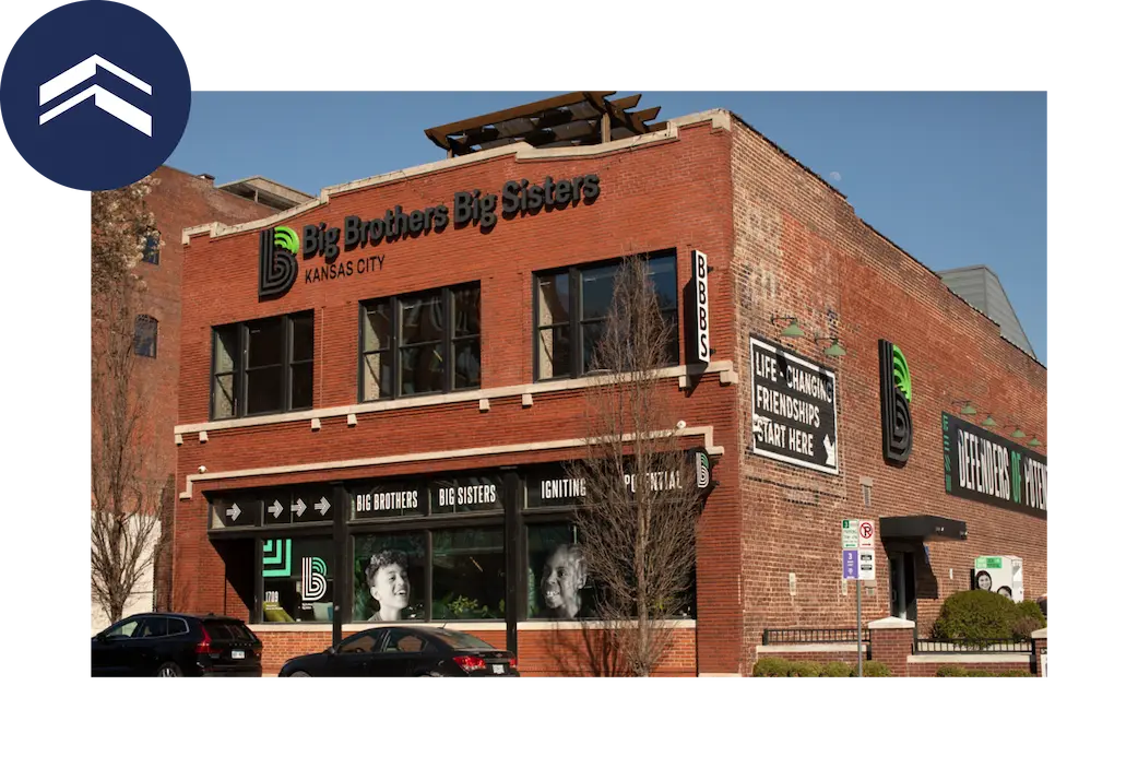 Exterior view of a two-story brick building for Big Brothers Big Sisters Kansas City, showing signage, large black-and-white photos of children in the windows, and two parked cars in front on a sunny day.
