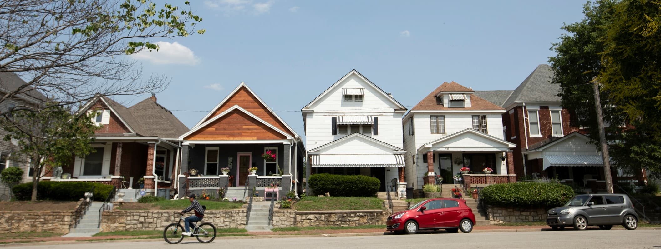 Row of small single-family houses along a typical Kansas City residential street, with a cyclist riding past parked cars on a sunny day.